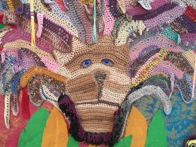 De afbeelding toont een kleurrijke muurkunstinstallatie gemaakt van textiel en haakwerk, vaak aangeduid als 'yarn bombing' of straatkunst. 