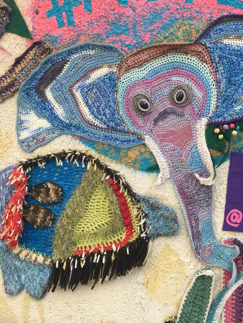 De afbeelding toont een kleurrijke muurkunstinstallatie gemaakt van textiel en haakwerk, vaak aangeduid als 'yarn bombing' of straatkunst. 