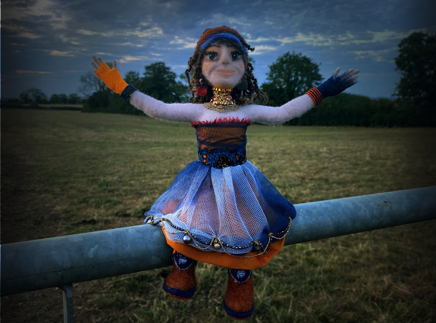 theatrical glitzy needle felted art doll sitting on a farm gate