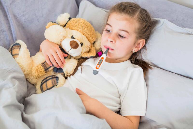 Kleines Mädchen liegt krank unter einer Decke im Bett. Sie hat Ihren Teddyim Arm und ein Fieberthermometer im Mund.
