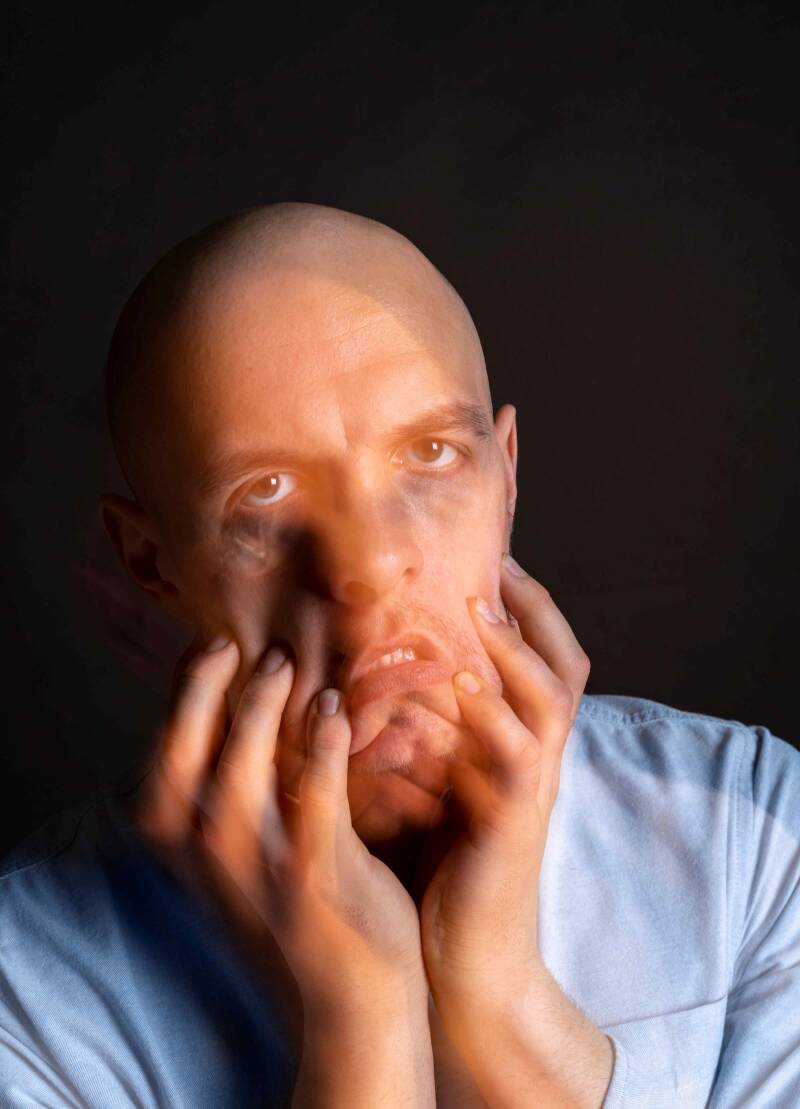 Ein Mann vor schwarzem Hintergrund greift sich ins Gesicht und zieht die Wangen nach unten. Dies soll sein Leiden in Verbindung mit seiner Schizophrenie veranschaulichen.