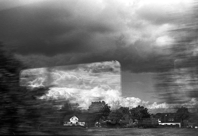 Analoge zwart wit foto gemaakt met een 35mm kleinbeeld camera; Noord Duitsland, foto gemaakt vanuit een rijdende trein: landschap, wolken, huizen en de reflectie in het raam van de trein; veel beweging