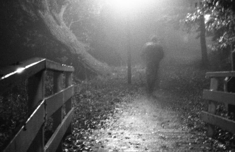 Analoge zwart wit foto gemaakt met een 35mm kleinbeeld camera; vaag silhouet van een man die 's avonds onder een lantarenpaal loopt; op de voorgrond een houten bruggetje, verder een paar bomen, het is mistig, de foto is onscherp en erg korrelig, poetisch