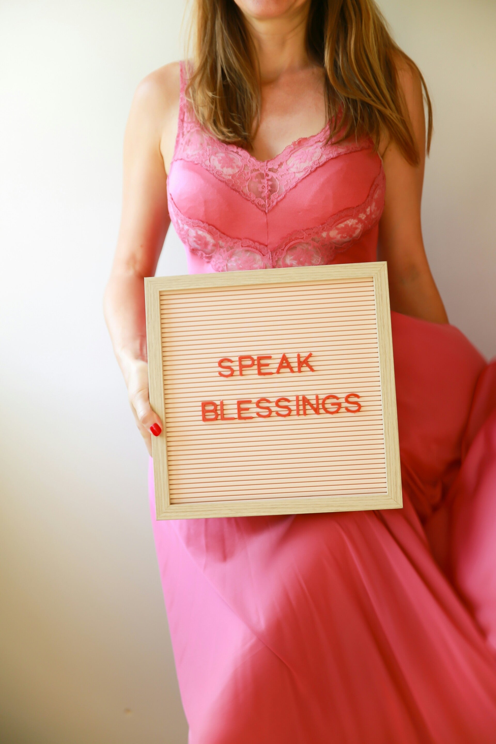 Person holding a letter board displaying the words “SPEAK BLESSINGS,” wearing a bright pink dress.