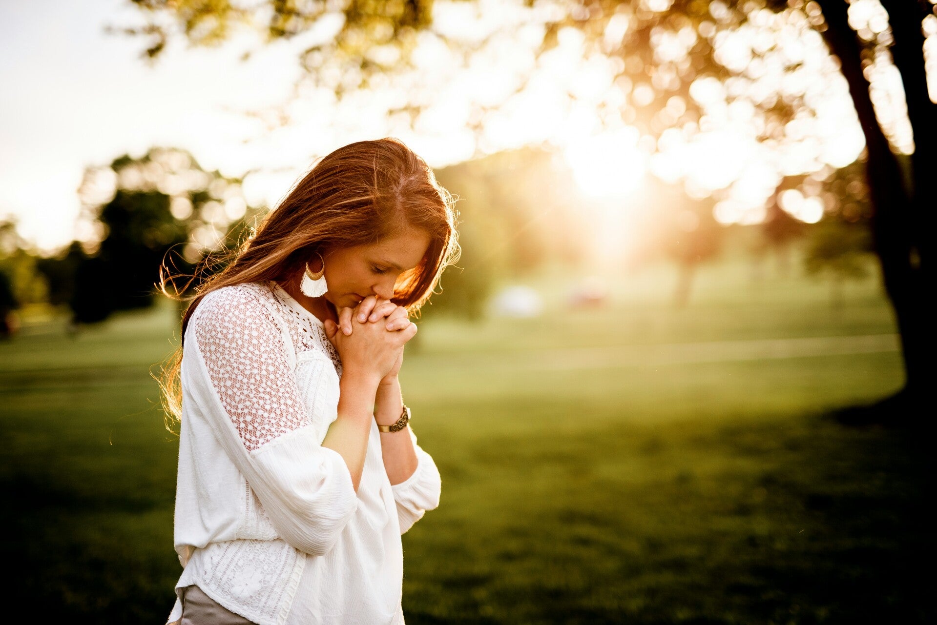 Woman standing outdoors at sunset with hands clasped and head bowed in prayer.