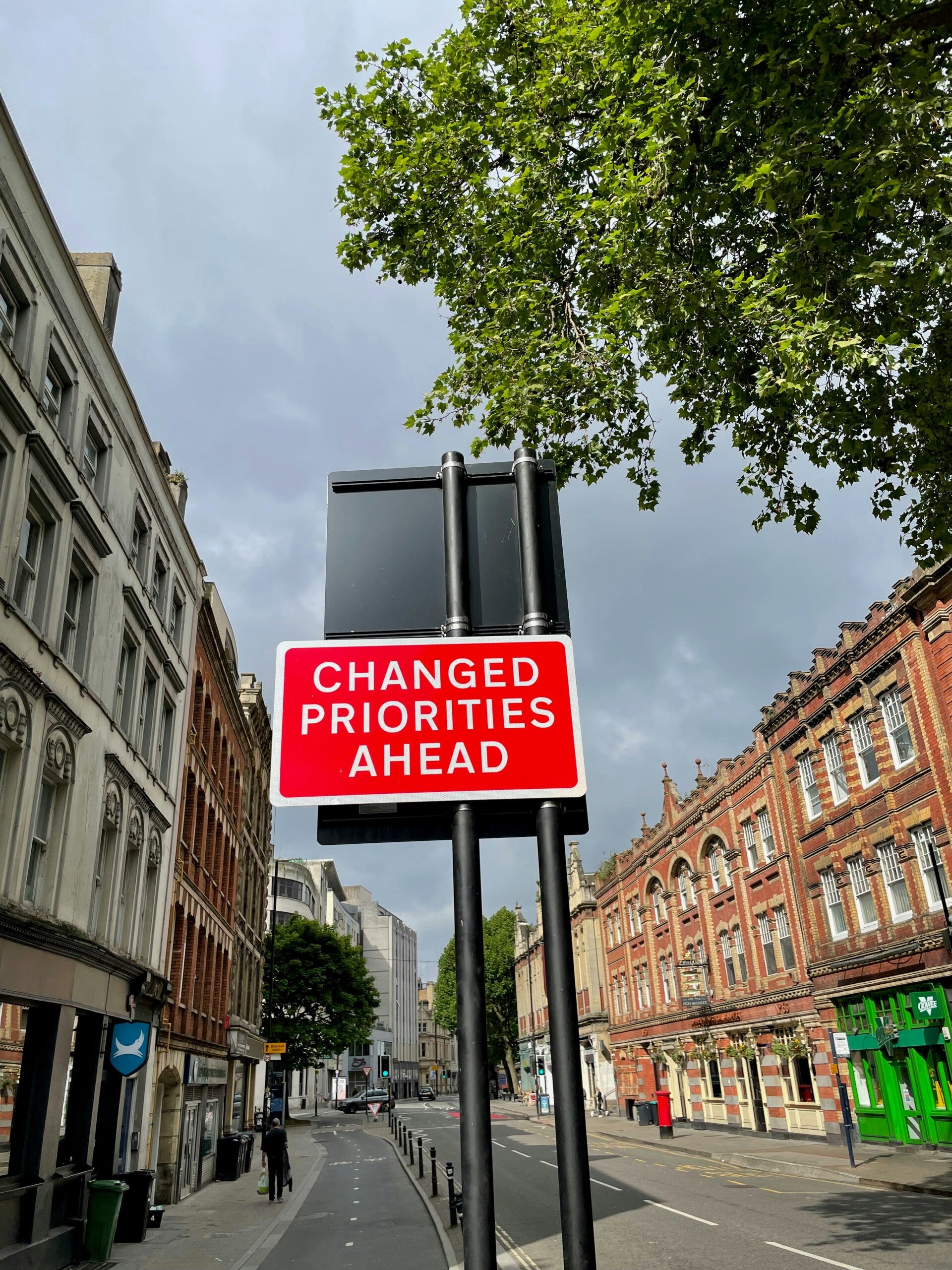 Red street sign reading ‘Changed Priorities Ahead’ in urban setting.