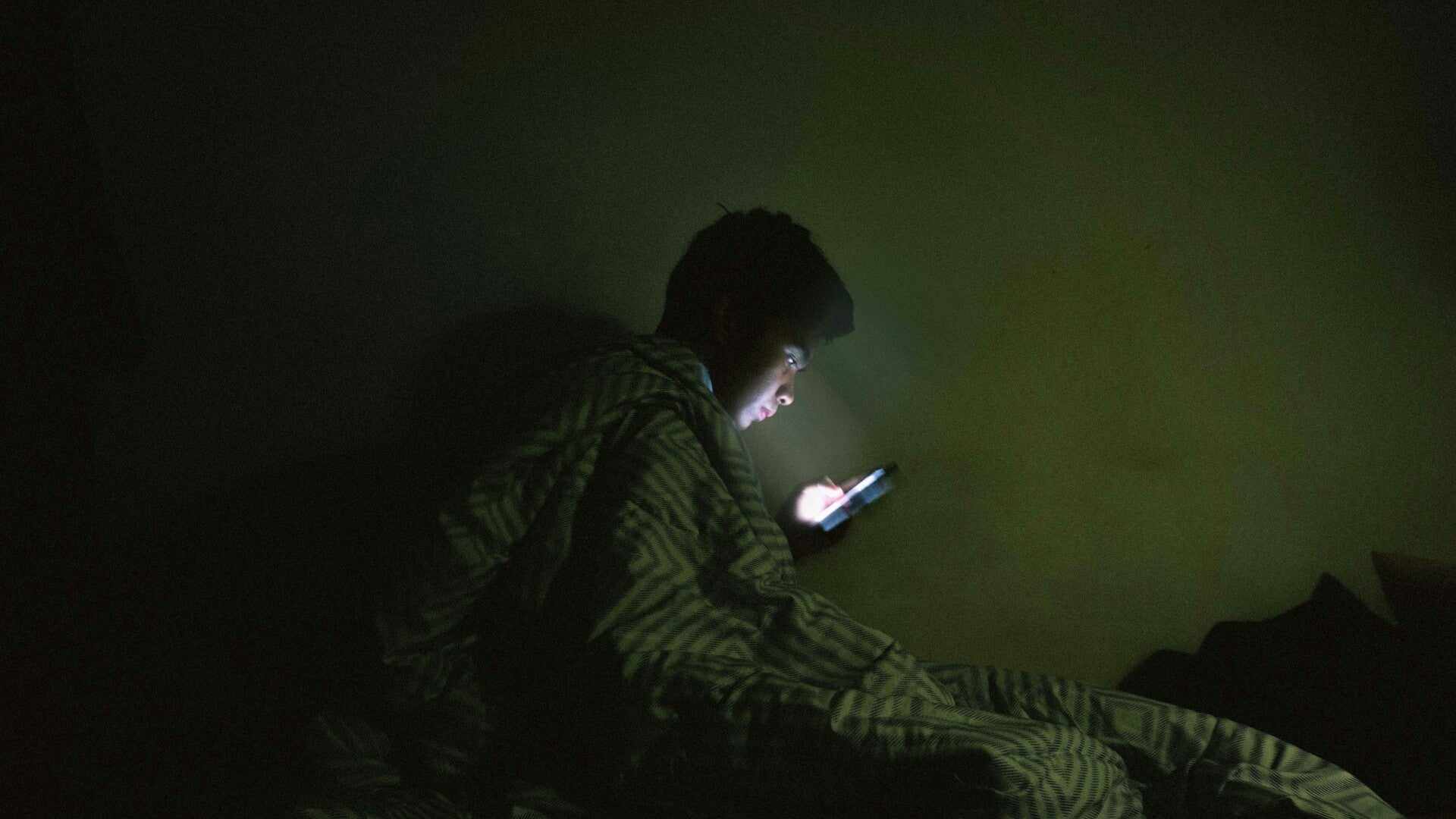 A young man in dark room. sitting in bed, wrapped in his blanket and face lit by phone. 