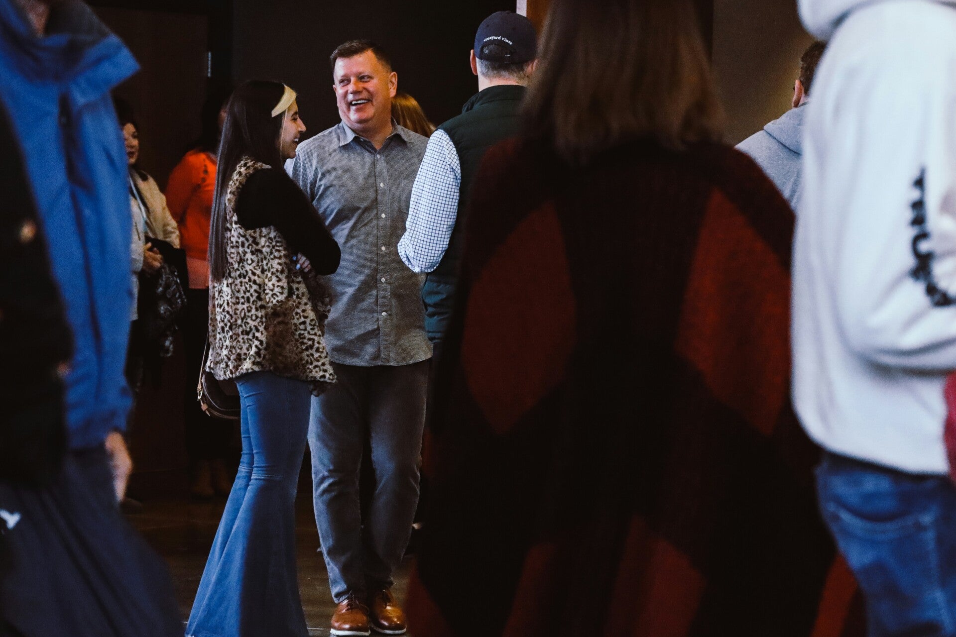 A man in a grey shirt laughing while talking to a young woman in a leopard print vest at a gathering.