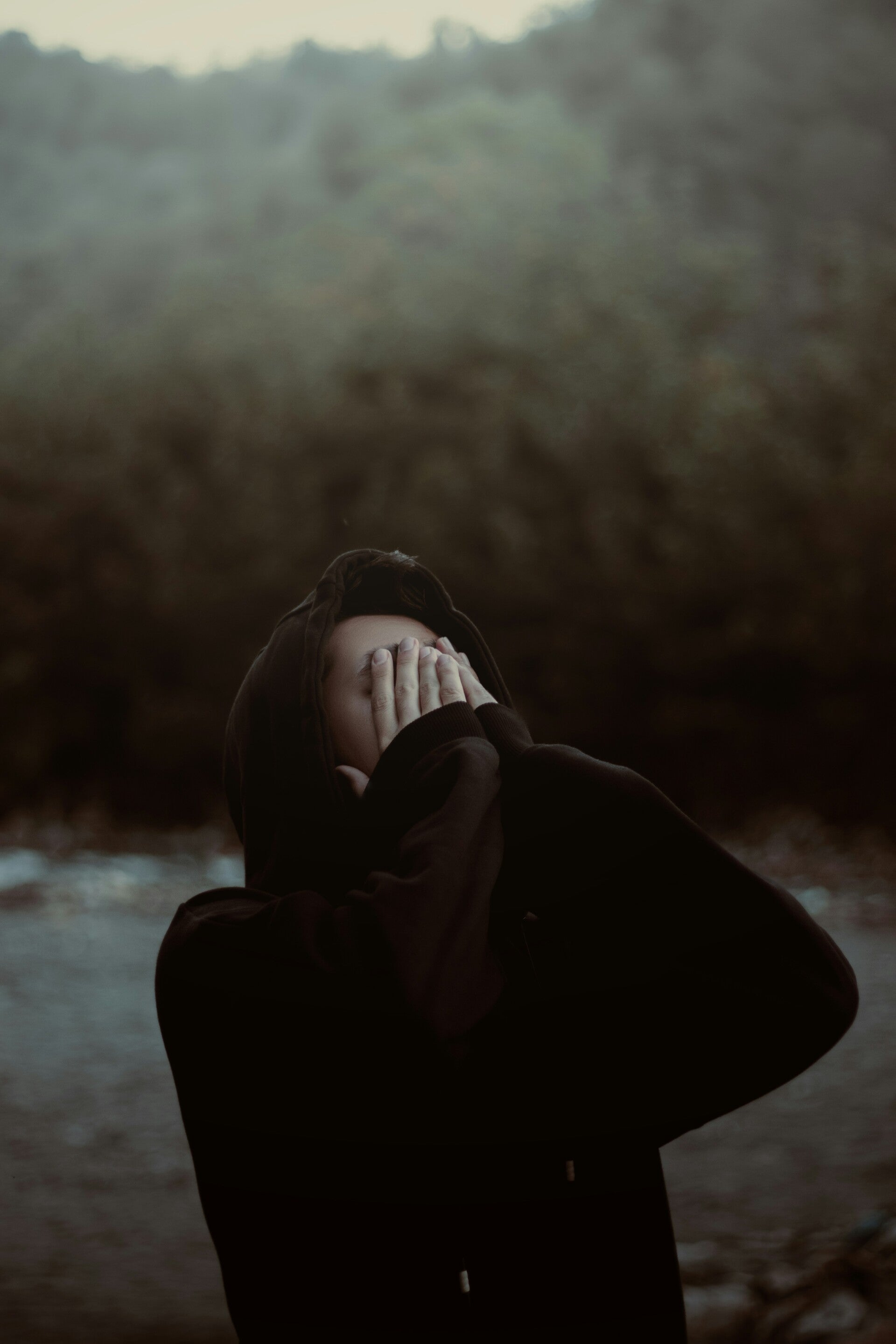 Person wearing a dark hoodie stands outdoors on unpaved ground, covering their face with both hands.