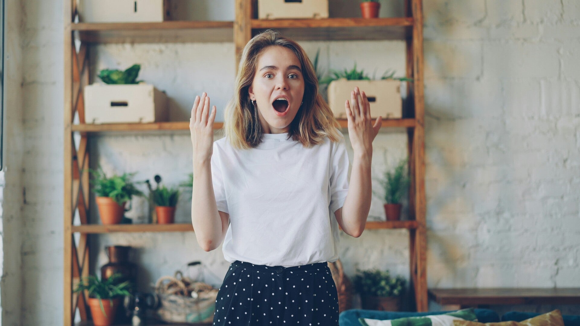 A woman reacts with surprise, hands raised and mouth open, standing in front of shelves with plants.