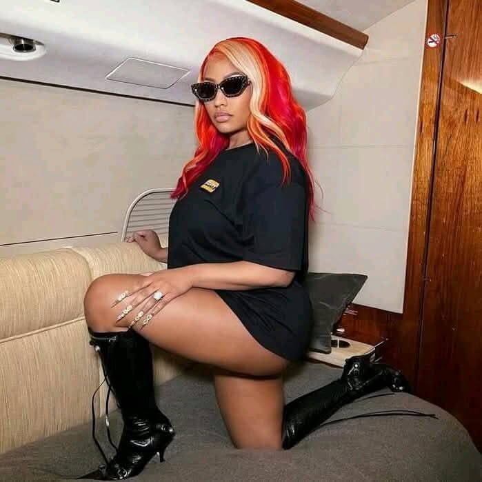 Nicki Minaj posing on a jet in a black t-shirt dress, black boots, shades, and red & blond hair. Looking confident and powerful.