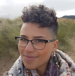 Close-up image of a light brown skinned female wearing black rimmed glasses. She has short curly hair on top and shaved at the sides. She is wearing a patterned scarf and smiling with her mouth closed. Behind her there are sand dunes and a cloudy sky.