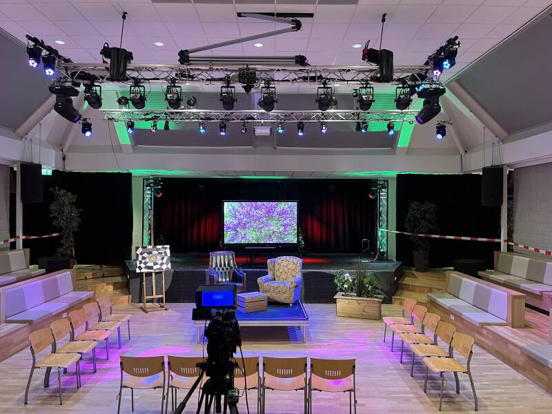 Uitgelichte zaal met daarin speakers en een klein podiumpje in het midden. Camera filmt het podiium