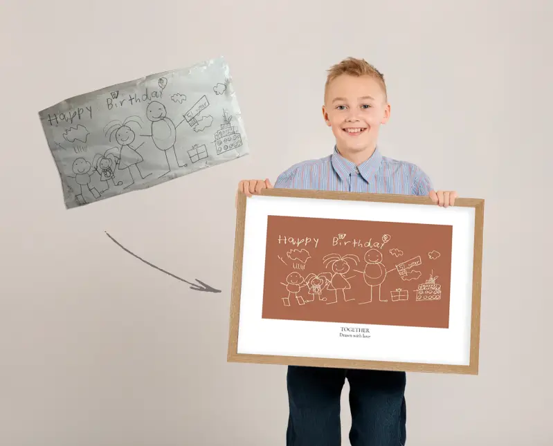 Smiling child holding a framed digital transformation of their original birthday family drawing.
