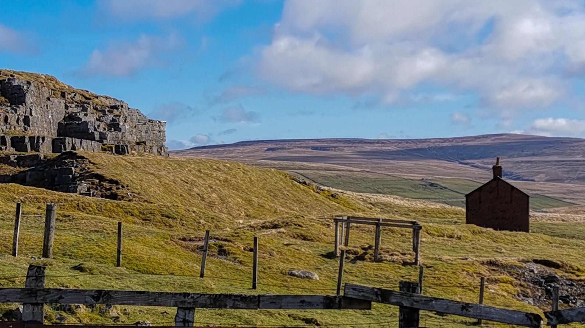 Landscape view across East Cow Green Mine showing scattered workings, a fenced shaft, quarry remains, and the red brick building in Upper Teesdale