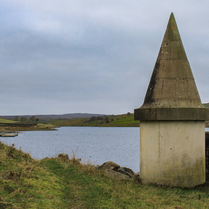 Stone obelisk marker beside Grassholme Reservoir with open water and pastoral landscape in the background, Teesdale