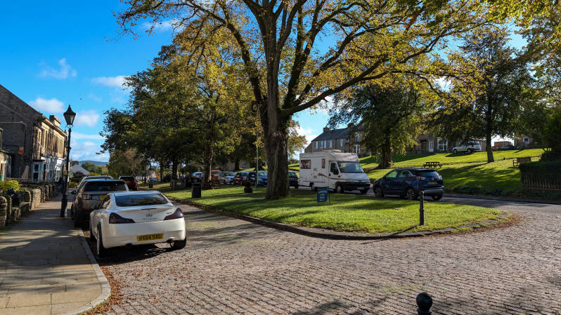 View of Horsemarket in Middleton-in-Teesdale, with its tree-lined village green, traditional stone buildings and roadside parking in County Durham.