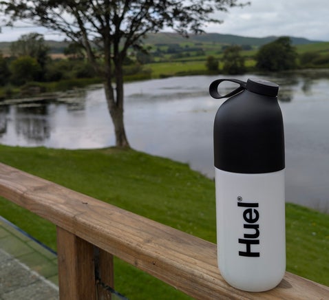 Huel shaker bottle placed beside a scenic lake during an outdoor adventure