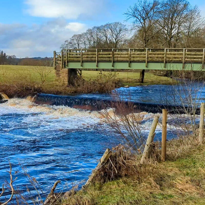Footbridge crossing the River Greta beside the weir near Bowes, marking the riverside section of the God's Bridge walk.