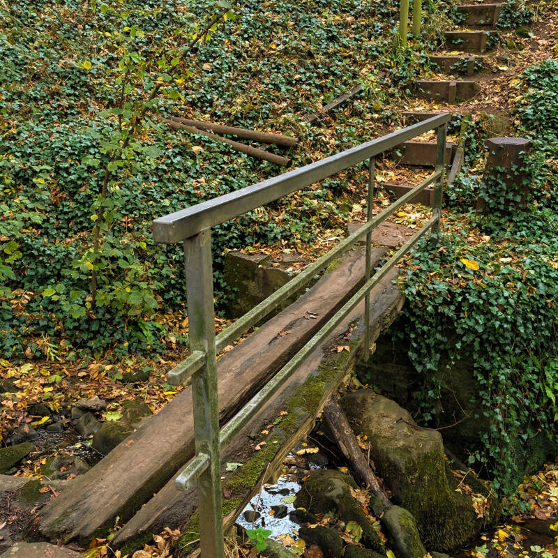 A narrow wooden footbridge crossing a rocky beck below Sledwich Hall, with woodland steps leading uphill.