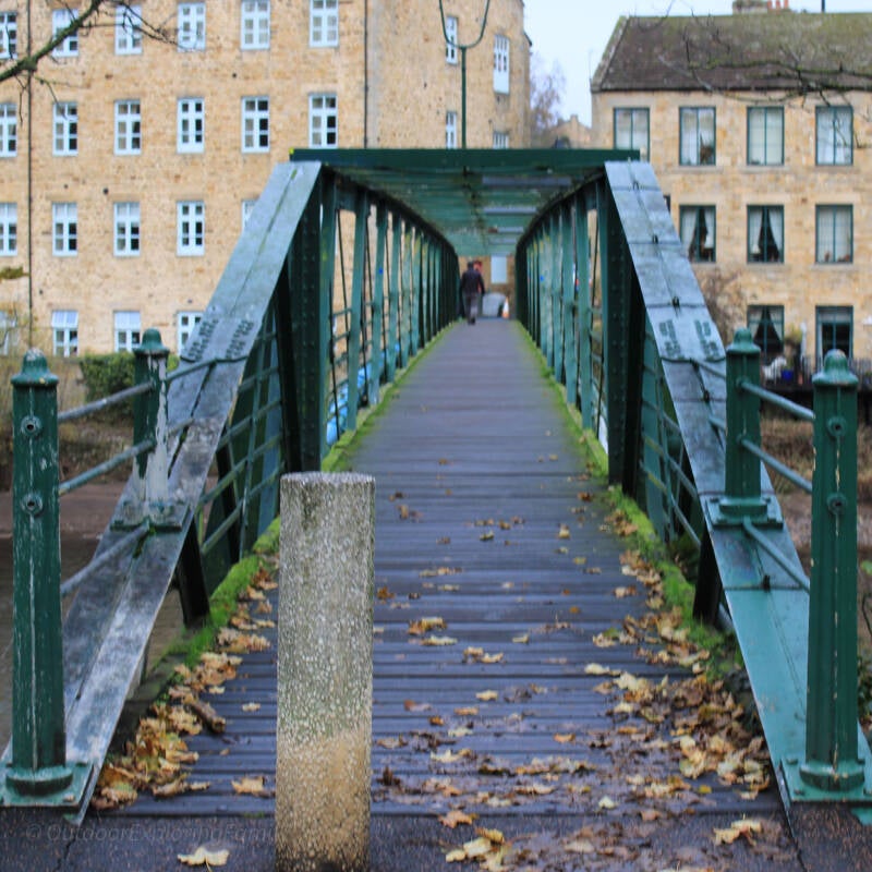 View of Thorngate Bridge from the riverside path, showing the green iron footbridge and old mill buildings beside the River Tees.