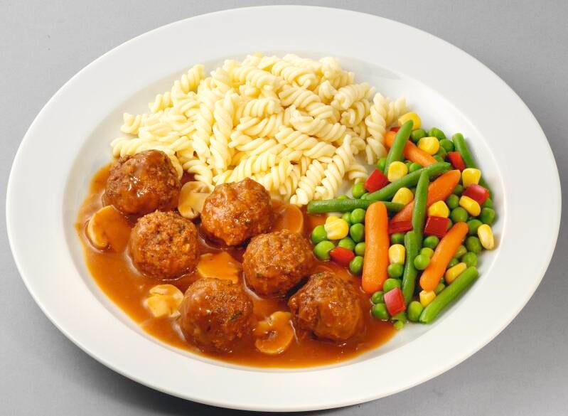 Ein weißer Teller mit Hackbällchen in brauner Soße mit Pilzen, dazu Spiralnudeln und buntes Gemüse aus Möhren, Erbsen, Mais, Bohnen und Paprika.