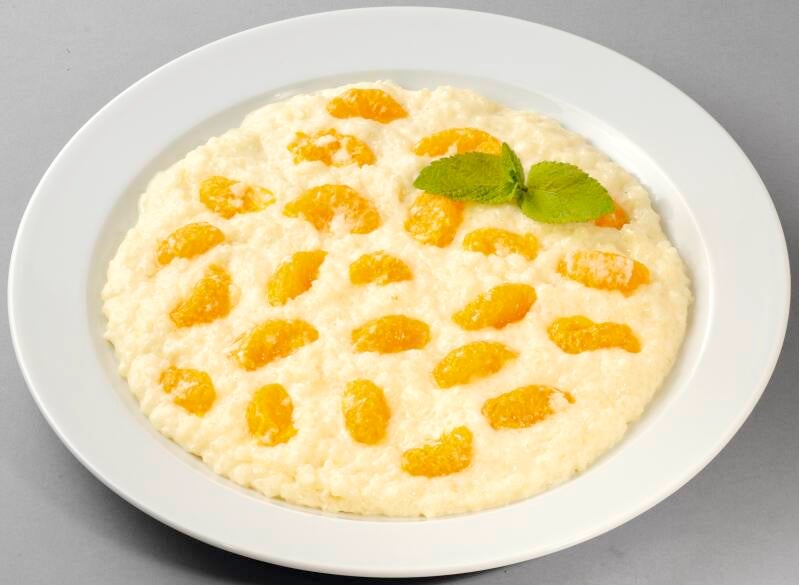 Ein weißer Teller mit Milchreis, belegt mit Mandarinenstücken. Obenauf liegt ein Zweig frische Minze zur Dekoration.
