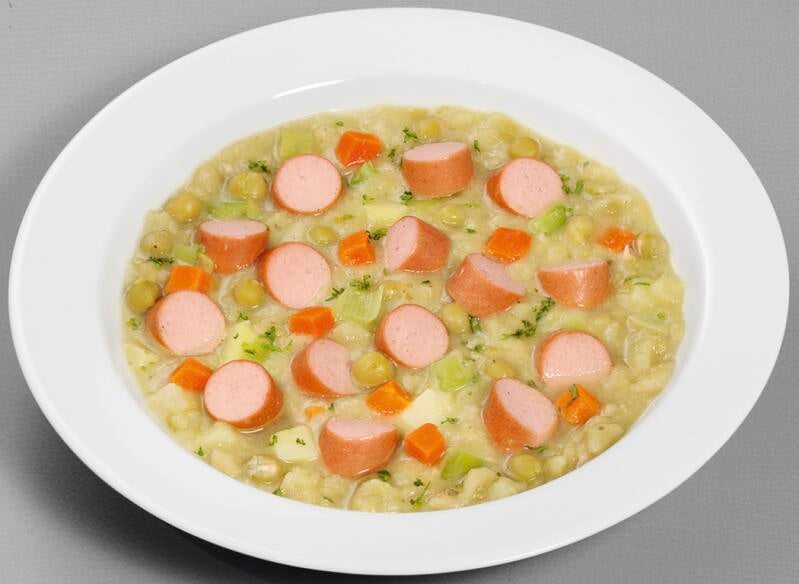 Ein weißer Teller mit einem Eintopf aus Erbsen, Kartoffeln, Karotten und Lauch, garniert mit in Scheiben geschnittenen Würstchen.
