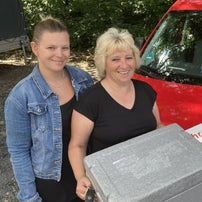 Zwei Mitarbeiterinnen vom Menüservice mit Herz stehen lächelnd vor einem roten Lieferwagen mit Firmenlogo und halten eine Menübox in der Hand.