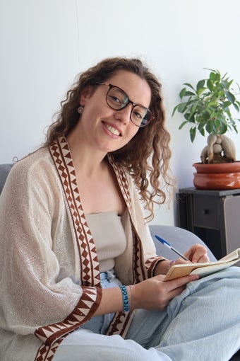 Retrato de una mujer joven, sonriente, con gafas. Sentada en un sofá con las piernas cruzadas y escribiendo en un cuaderno con un bolígrafo azul