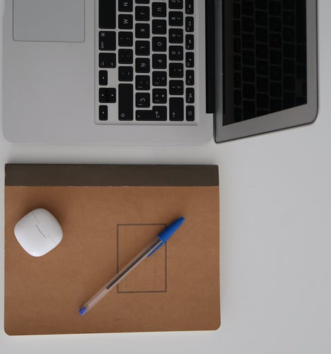Escritorio con una laptob, un cuaderno marrón, un bolígrado azul y una funda blanca de auriculares