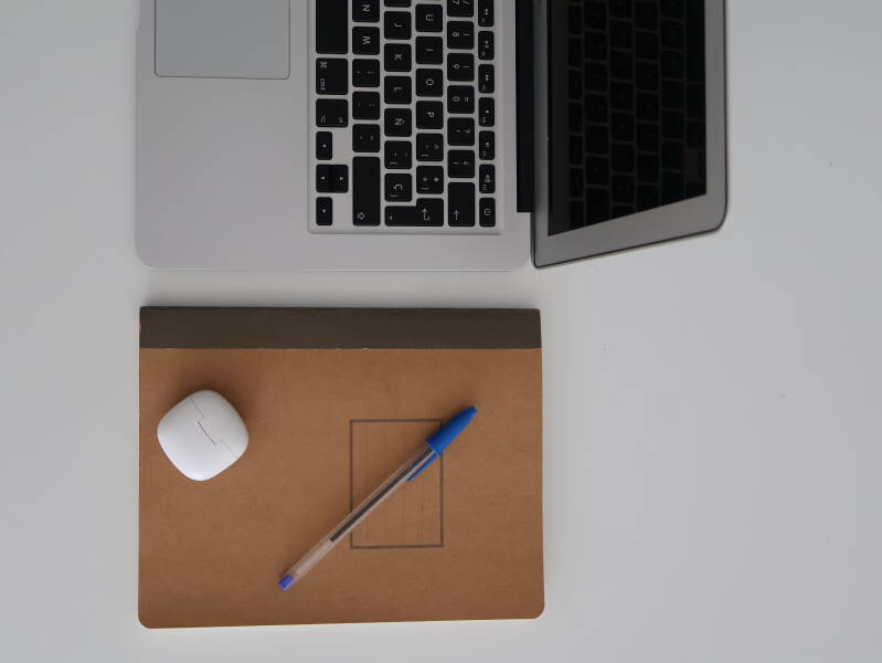 Escritorio con una laptob, un cuaderno marrón, un bolígrado azul y una funda blanca de auriculares