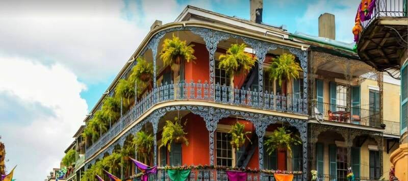 Colorful French Quarter building in New Orleans with iron lace balconies, hanging ferns, and Mardi Gras flags. Historic architecture on Royal Street with red facade and blue trim.