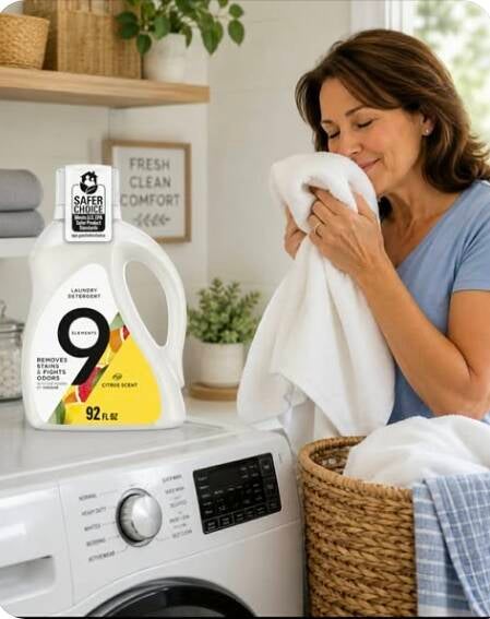 Woman enjoying fresh clean laundry washed with 9 Elements Citrus Liquid Laundry Detergent. EPA Safer Choice, dye-free bottle sits on washing machine next to wicker basket. Bright laundry room with "Fresh Clean Comfort" decor.