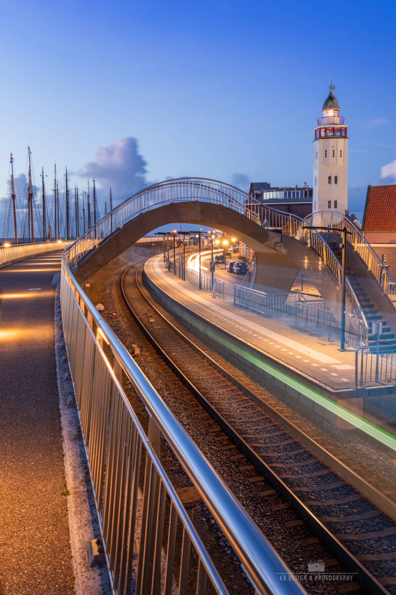 Sfeervolle avondfoto van Harlingen met de karakteristieke vuurtoren, spoorbrug en een voorbijrijdende trein met prachtige lichtstrepen tijdens het blauwe uur.
