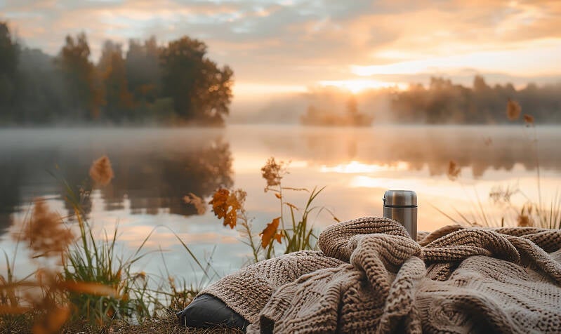 blanket and mug near lake at sunset tress in background