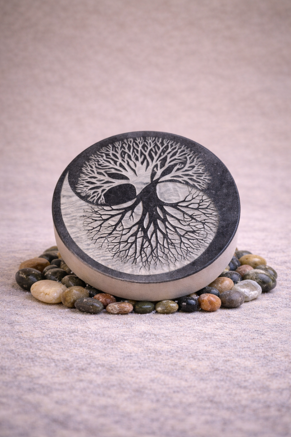 Galet rond en pierre naturelle gravé de l’Arbre de Vie, posé sur un lit de petits galets, sur fond neutre.