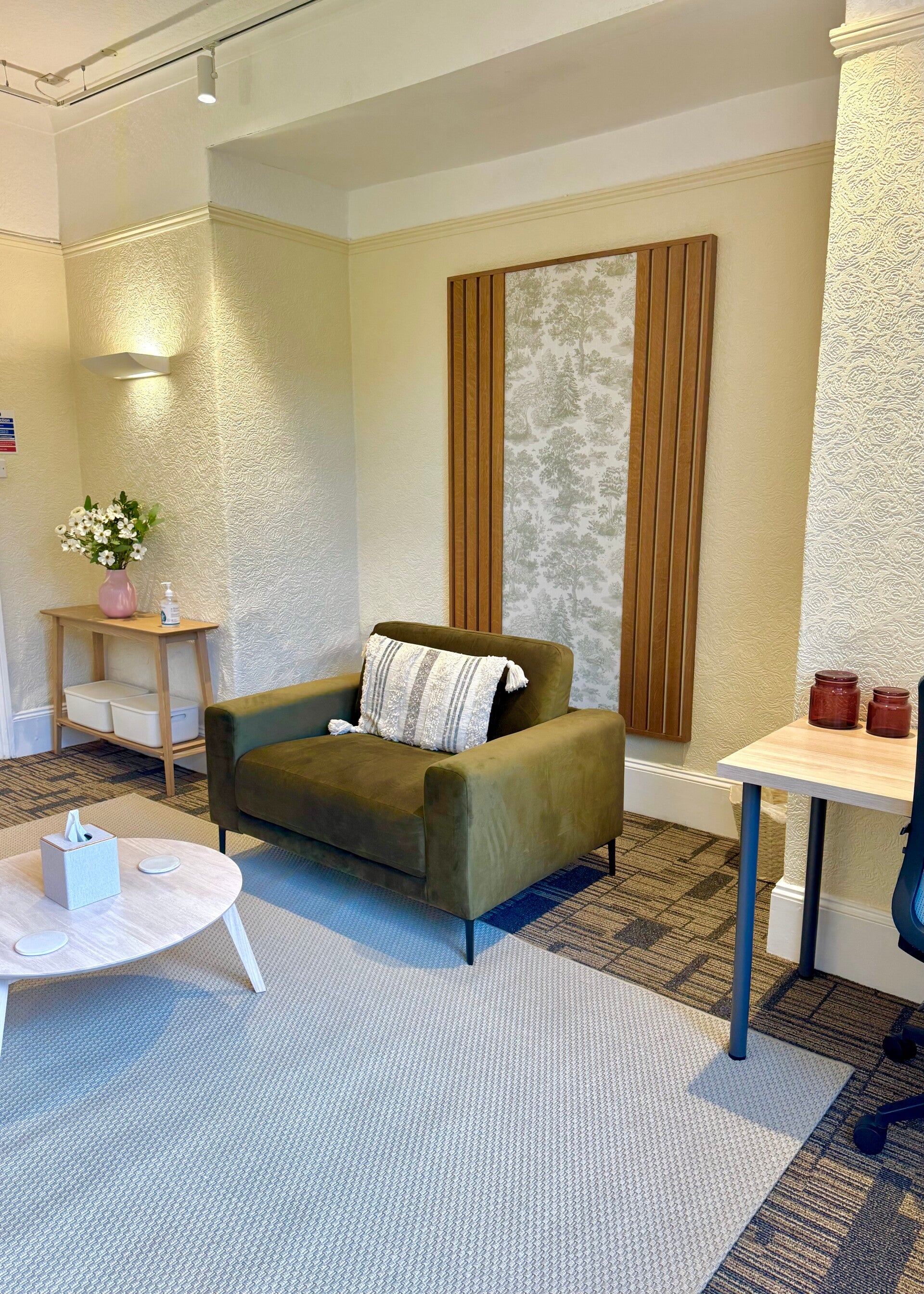 In the foreground, the corner of a desk. In the midground a green armchair and coffee table in front of a forest themed wall decal. In the background, a console table with vase and flower arrangement.