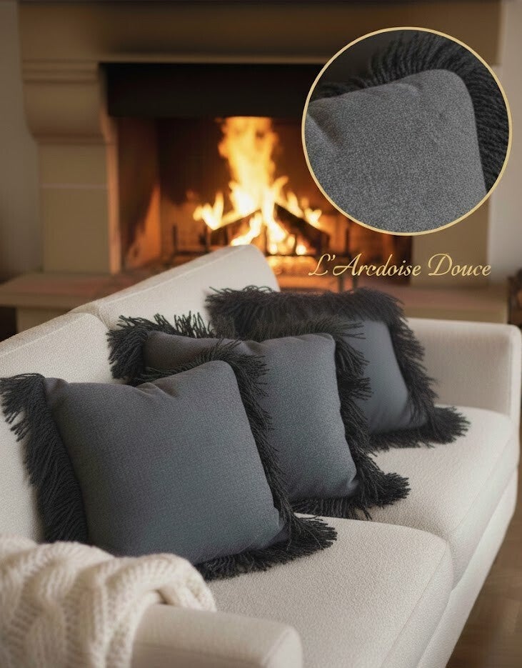 Trois coussins artisanaux gris à franges disposés sur un canapé blanc dans un salon chaleureux avec feu de cheminée. Artisanat Bordeaux
