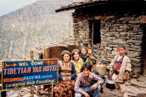 Photo of Maya Frost, founder of Collapse Forward, sitting in front of a house made of stacked stones, sitting with a group of young Nepalese people, with a sign that says: ¨Wel come to Tibetian Yak Hotel, Lodging and Fooding¨ in 1982