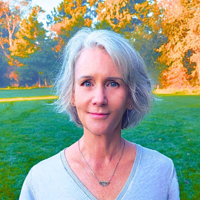 Maya Frost, founder of Collapse Forward, standing in a grassy area with colorful fall leaves on trees in background.