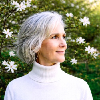 Maya Frost, founder of Collapse Forward, wearing a white turtleneck sweater, standing in front of blooming spring tree.