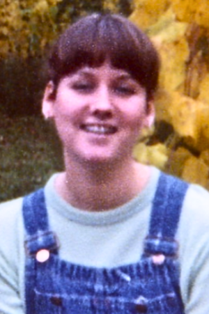 Photo of Maya Frost, founder of Collapse Forward, at age 17, wearing overalls and a grey sweatshirt underneath, background is yellow fall leaves.