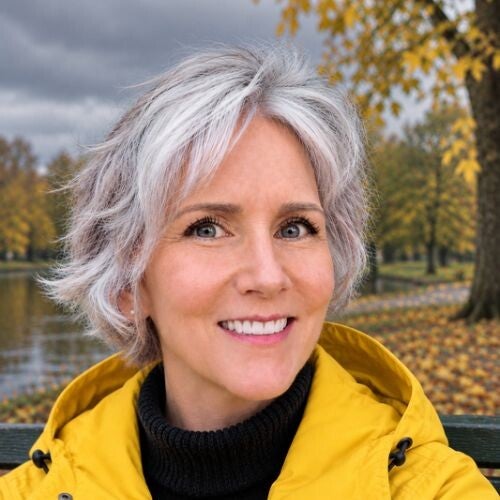 Maya Frost, creator of Lucid Longing, smiling, wearing a yellow long-sleeved shirt in a forest with green and yellow changing leaves.