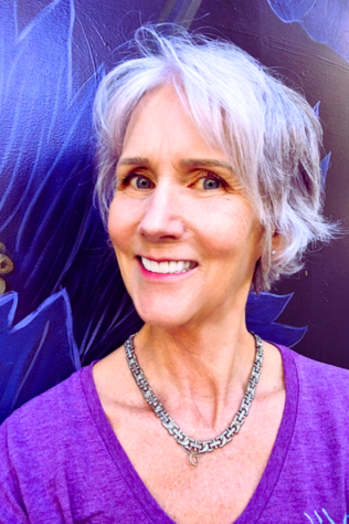 photo of Maya Frost, trauma-infomred breakthrough transformational coach, wearing a purple v-neck t-shirt against a wall mural in purple and blue