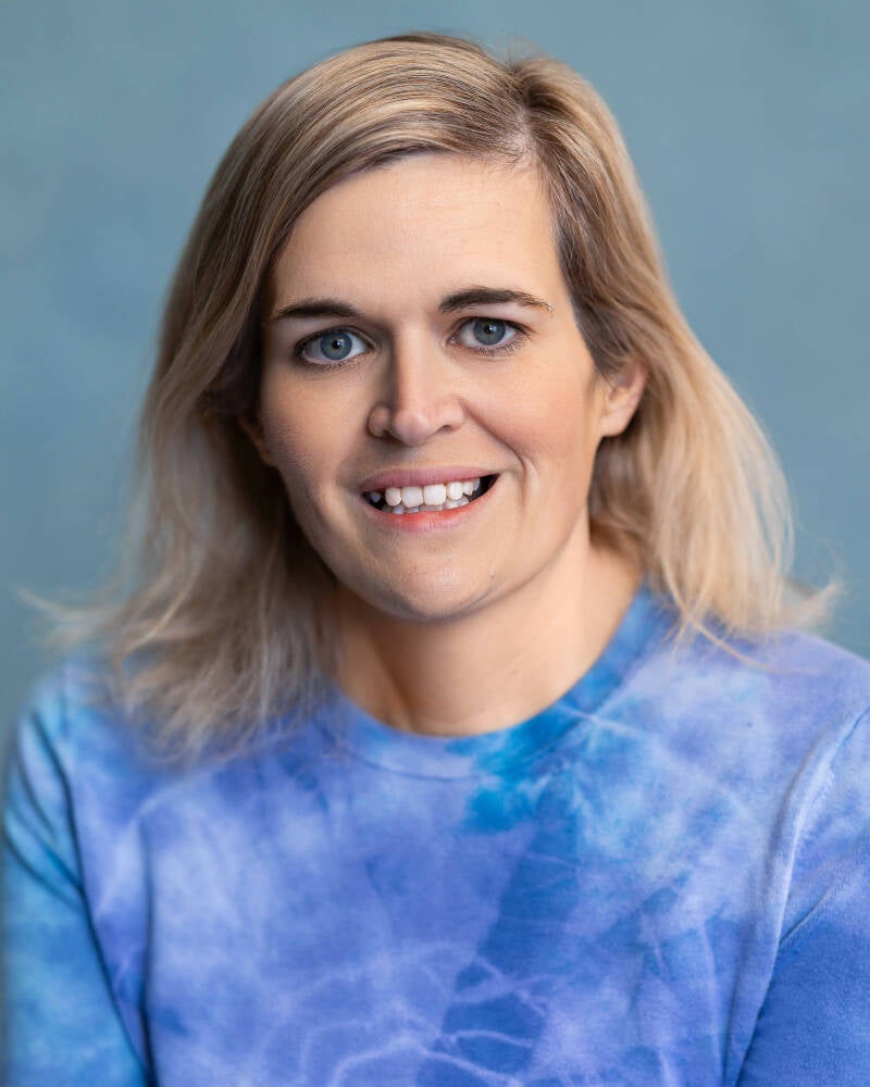A picture of a lady with blonde hair, blue eyes. Looks to be around late forties. She is posing for a professional headshot in studio setting. She is smiling and is wearing a blue tie dyed top.