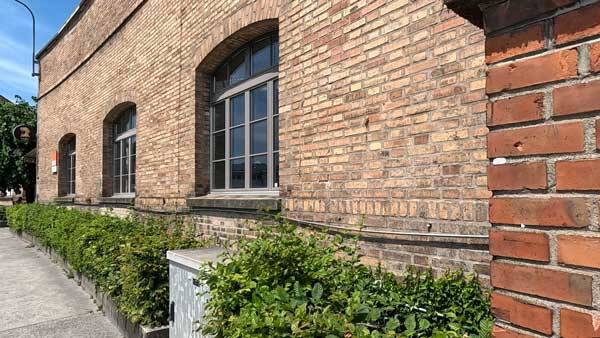 Nahaufnahme einer rustikalen Backsteinfassade mit bogenförmigen Fenstern und einer grünen Hecke im Vordergrund.