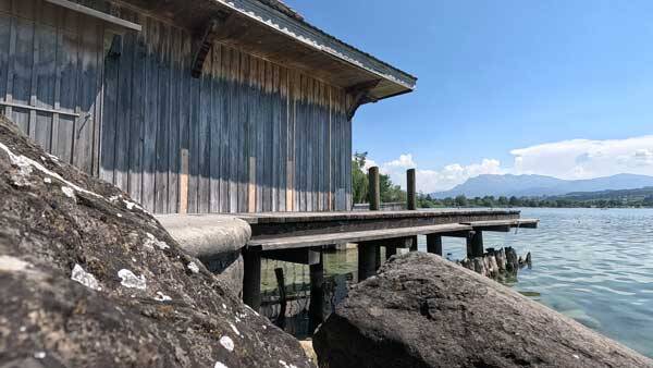 Ein historisches Bootshaus aus Holz am Ufer des Sempachersees, im Vordergrund grosse Steine und im Hintergrund eine Bergkette unter blauem Himmel.
