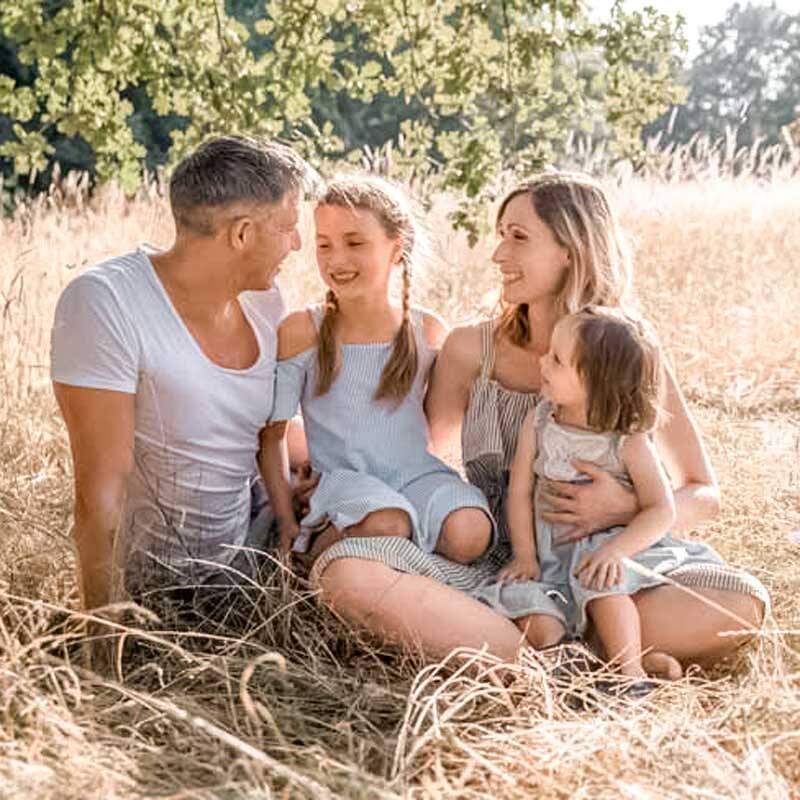 Familie sitzt im hohen, goldenen Gras unter einem Baum: Eltern und zwei Kinder lachen sich an und geniessen gemeinsam einen sonnigen Tag in der Natur