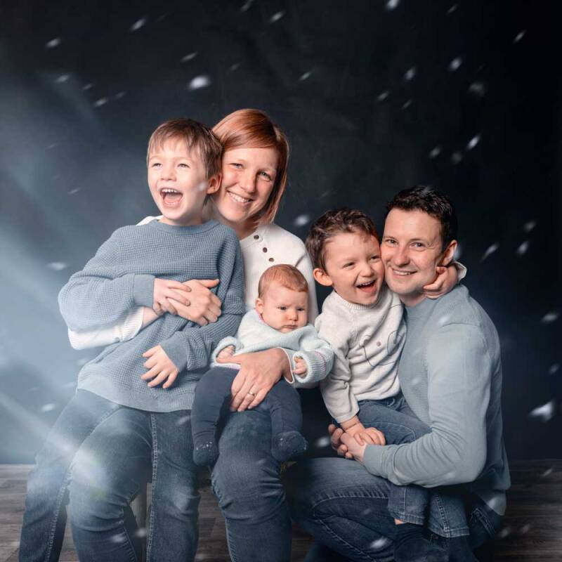 Ein fröhliches Familienfoto im Fotostudio mit Eltern und drei Kindern, die vor einem dunklen Hintergrund mit digitalem Schneeeffekt lachen.
